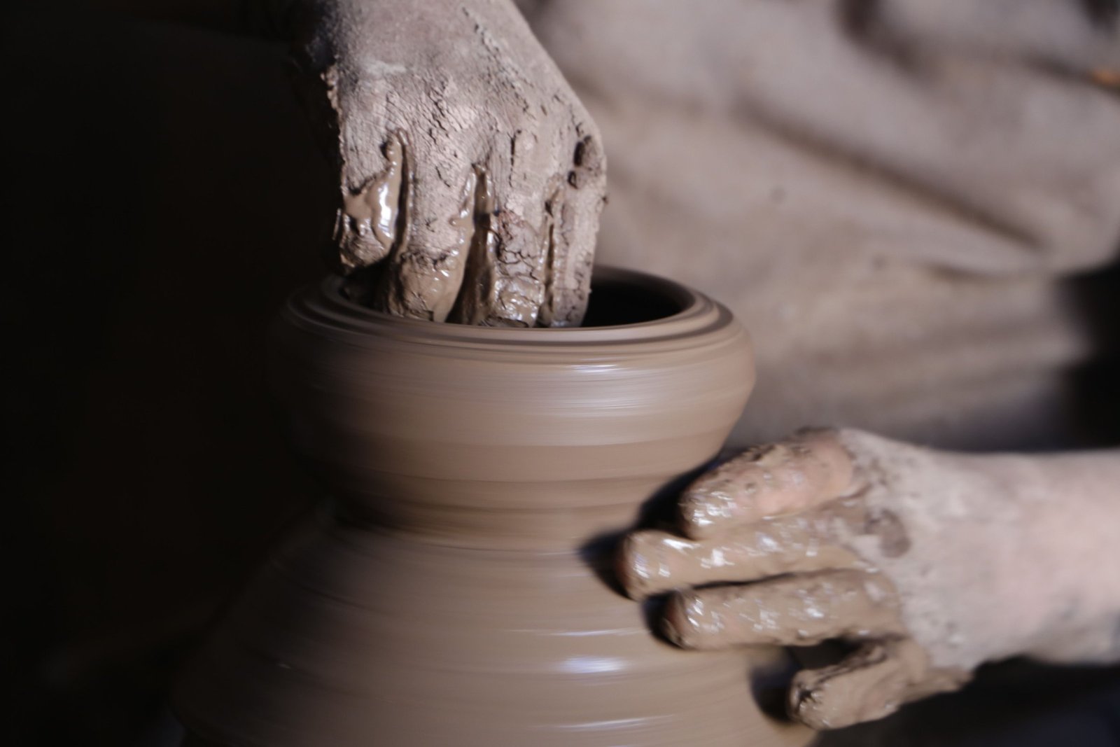 A potter's clay-covered hands shaping a vessel on the wheel